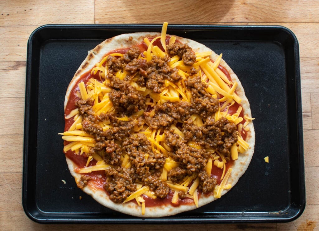 Ground beef that has been browned and seasoned with taco meat is sprinkled over cheddar cheese and tomato sauce on a piece of Naan bread before baking.