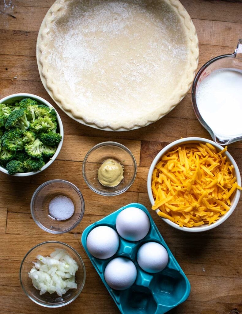 Ingredients to make a broccoli cheese quiche including cream, grated cheddar cheese, chopped broccoli and eggs.