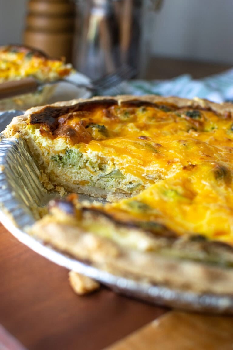 A broccoli and cheese quiche with one slice cut out and placed on a plate to show the inside that's loaded with chunks of broccoli and cheddar cheese.