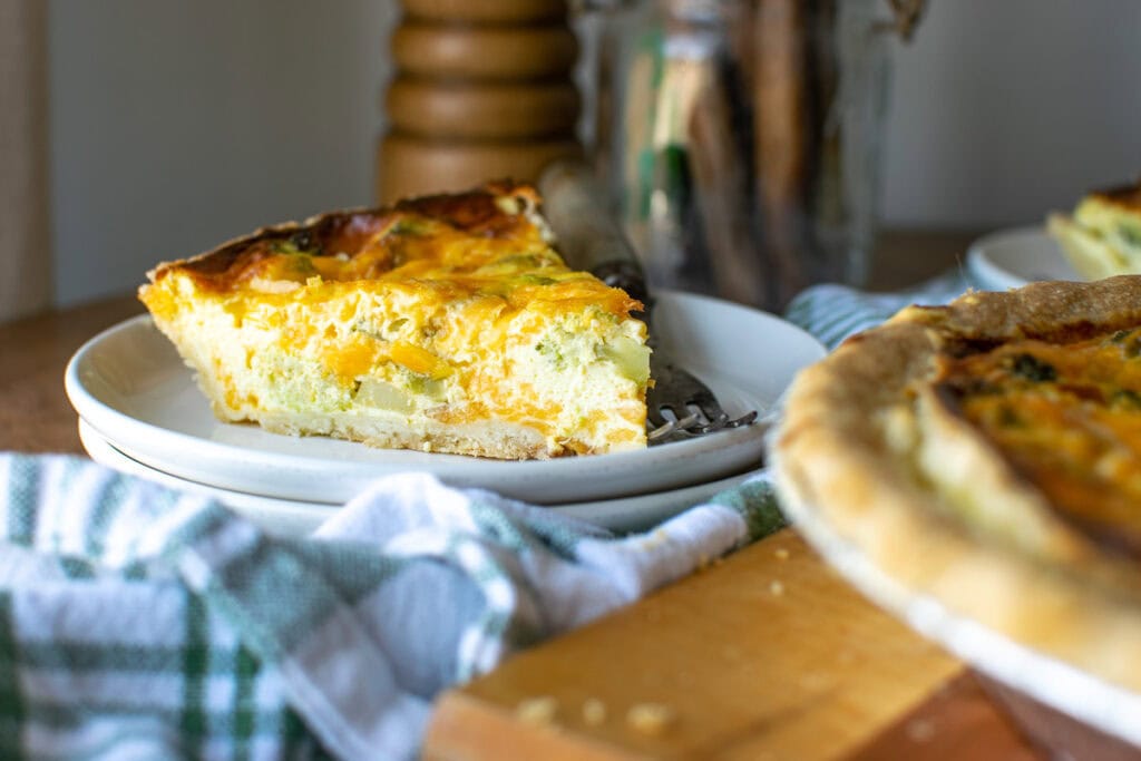 A slice of broccoli cheese quiche on a white plate with a green and white napkin underneath.