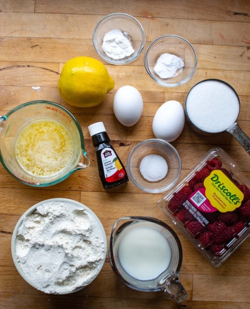 Ingredients to make lemon and raspberry muffins including sugar, lemons, fresh raspberries, flour and eggs.