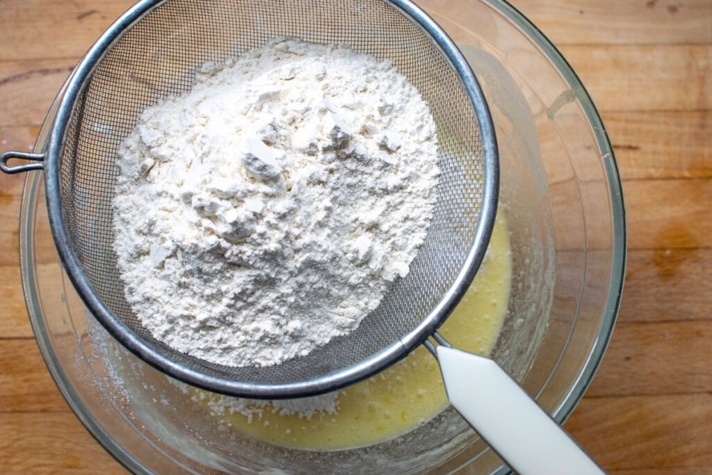 The dry ingredients for lemon raspberry muffins being sifted into the wet ingredients in a large mixing bowl.