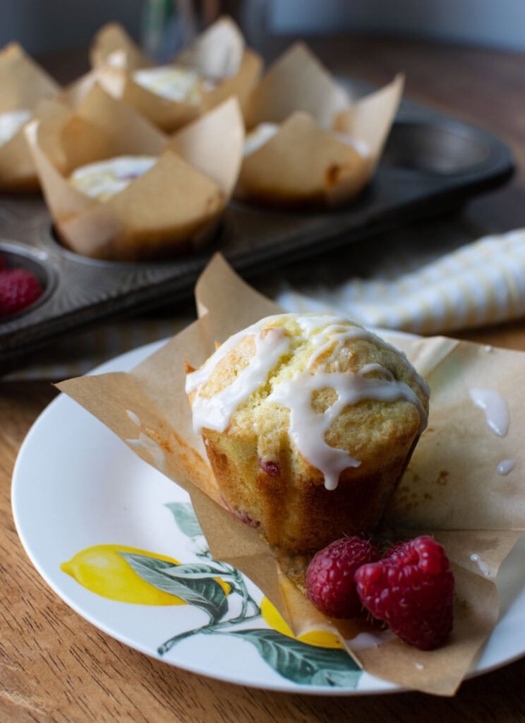 A lemon and raspberry muffin on a plate with images of lemons.  The parchment paper liner is pulled away from the muffin.