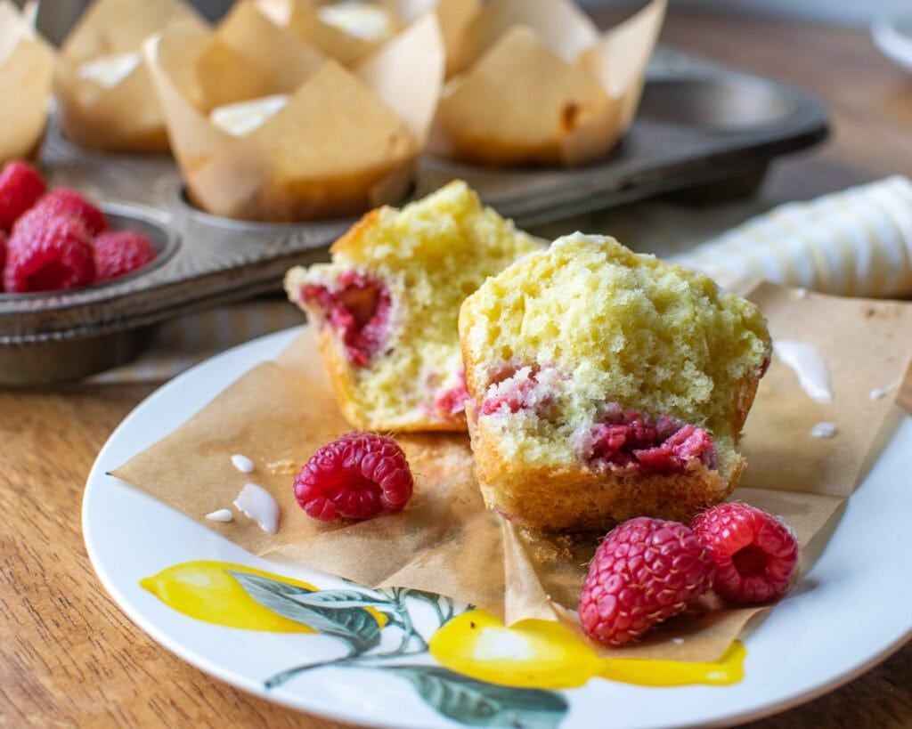 A lemon and raspberry muffin cut in half, on a plate decorated with a lemon pattern, and a few fresh raspberries sprinkled around.