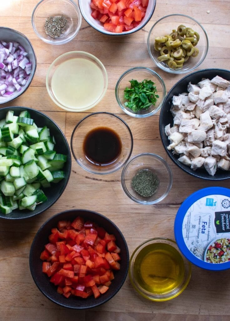 Ingredients to make a chicken Mediterranean salad including cucumber, tomato, olives, red bell pepper, red onion, and cooked chicken breast.