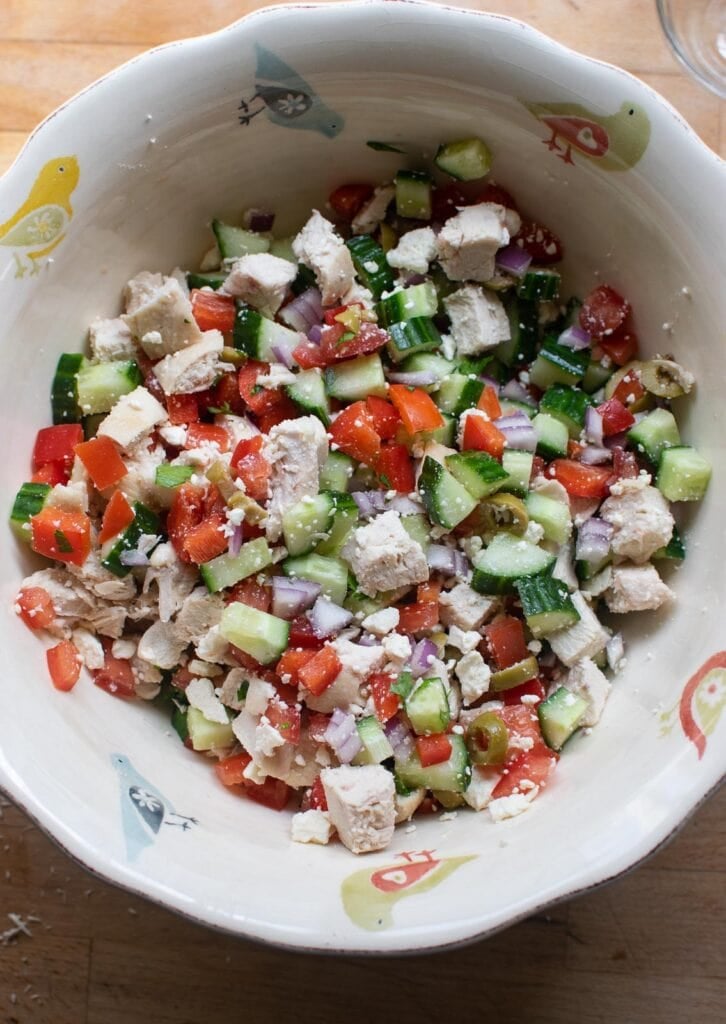 Ingredients for a mediterranean salad including chicken, feta cheese, diced cucumber, red bell pepper and red onion tossed together in a mixing bowl.