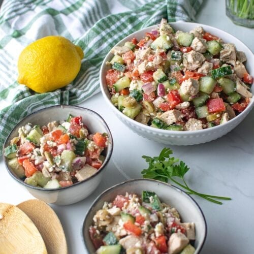 A serving bowl of mediterranean salad with chicken and feta with two small bowls full beside it. A lemon and parsley springs sit beside the bowls.
