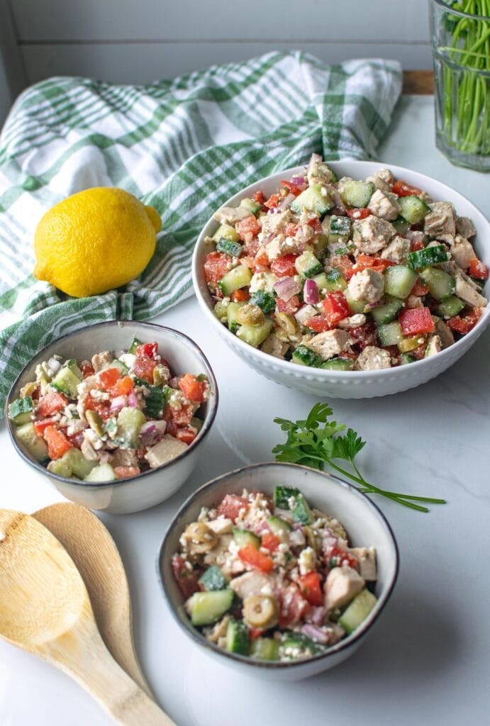 A serving bowl of mediterranean salad with chicken and feta with two small bowls full beside it.  A lemon and parsley springs sit beside the bowls.