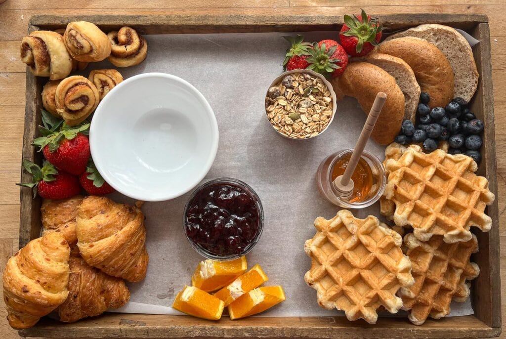 Breads elements are added to a charcuterie board, and bowls are filled with jams and granola, as well as some strawberries and sliced oranges.