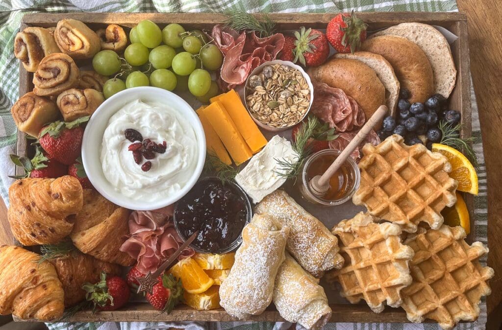 A completed brunch charcuterie board filled with bagels, chocolate and cheese croissants, yogurt, mini waffles, grapes and cheddar cheese.