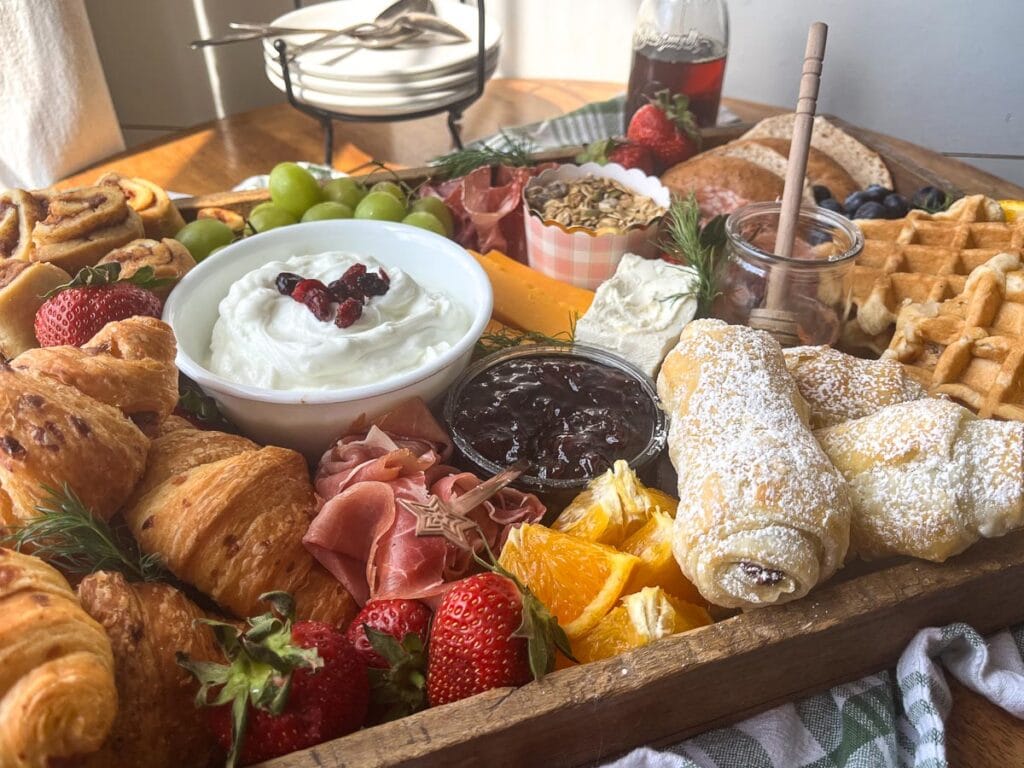 A brunch board with a variety of breads, pastries, yogurt, granola, deli meats and cheese.