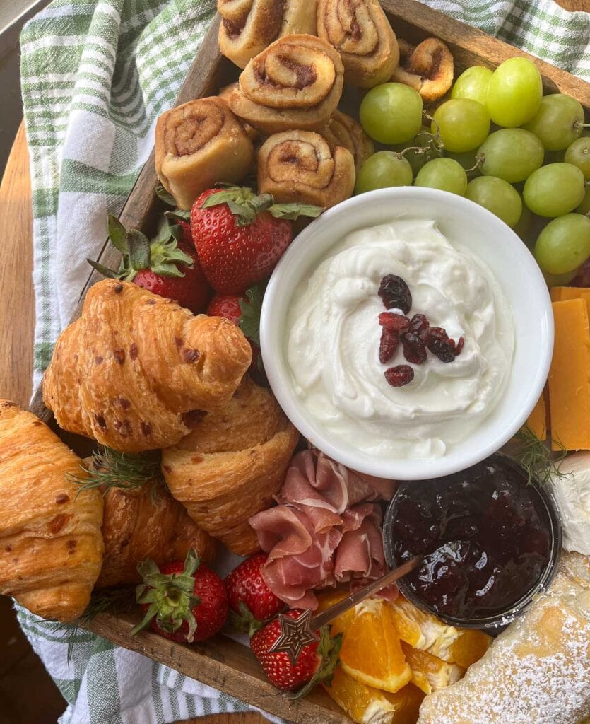 A brunch charcuterie board with mini croissants, a bowl of yogurt, cinnamon rolls, grapes and cured meats.