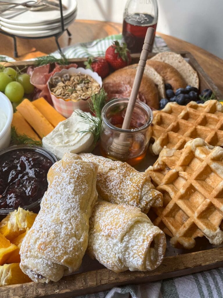 Pastries and breads on a brunch charcuterie board for spring, including chocolate croissants, mini Belgian waffles, sliced bagels, cream cheese, jam and fruit.