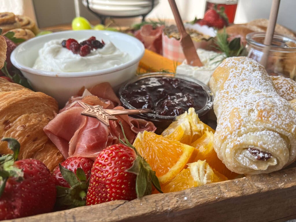 A close up of items on a brunch charcuterie board that includes chocolate croissants, sliced oranges, strawberries, yogurt, bagels, jam and cream cheese.