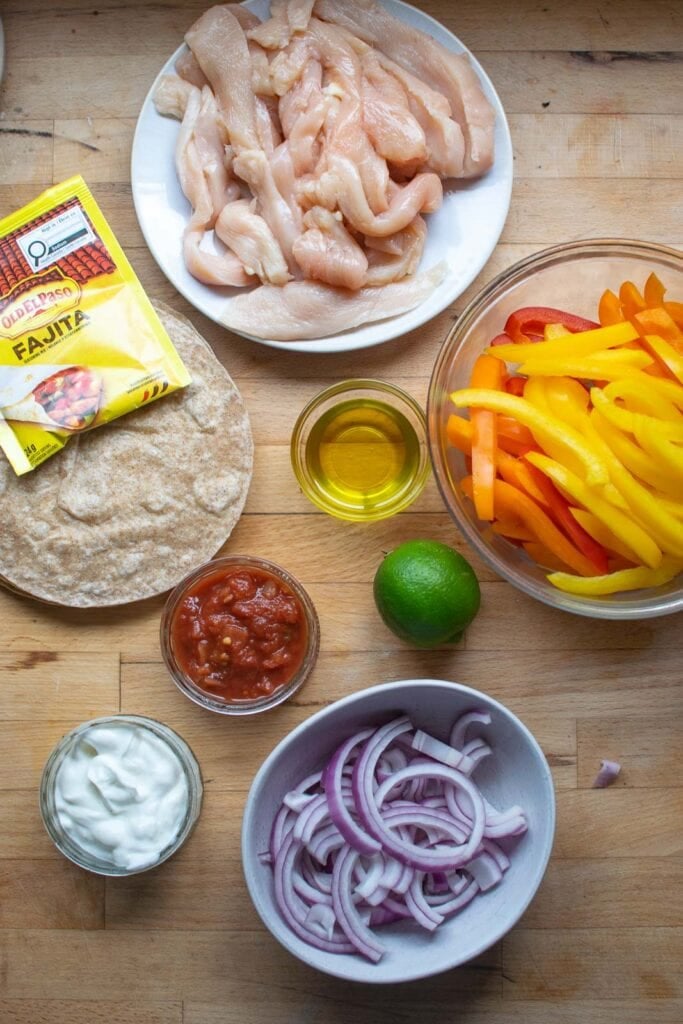 Ingredients to make chicken fajitas including  sliced chicken pieces, sliced bell peppers, red onion, fajita seasoning, sour cream and salsa.