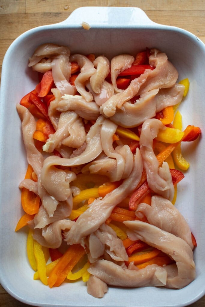 Raw sliced chicken on top of sliced bell peppers in a casserole dish
