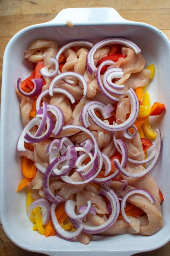 Raw sliced red onions on top of chicken pieces and sliced bell peppers in a casserole dish.