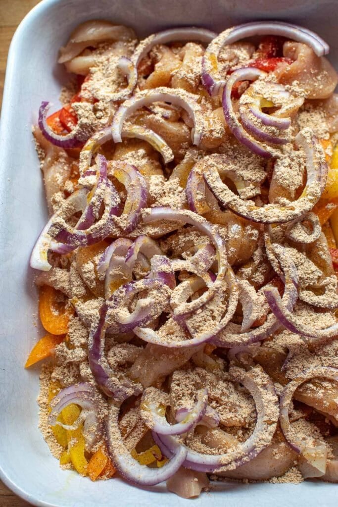 Raw chicken, bell peppers and red onions sprinkled with fajita seasoning in a casserole dish.