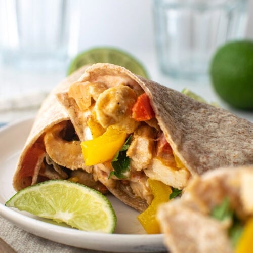 A oven baked chicken fajita wrap cut in half and served on a white plate with a lime wedge.