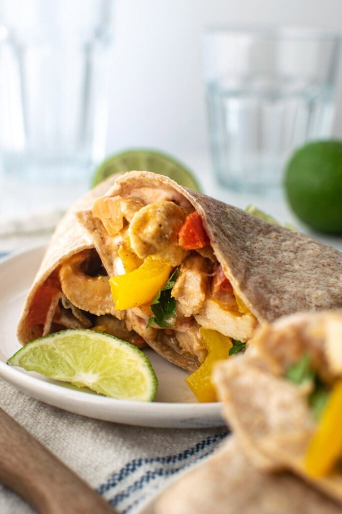 A oven baked chicken fajita wrap cut in half and served on a white plate with a lime wedge.