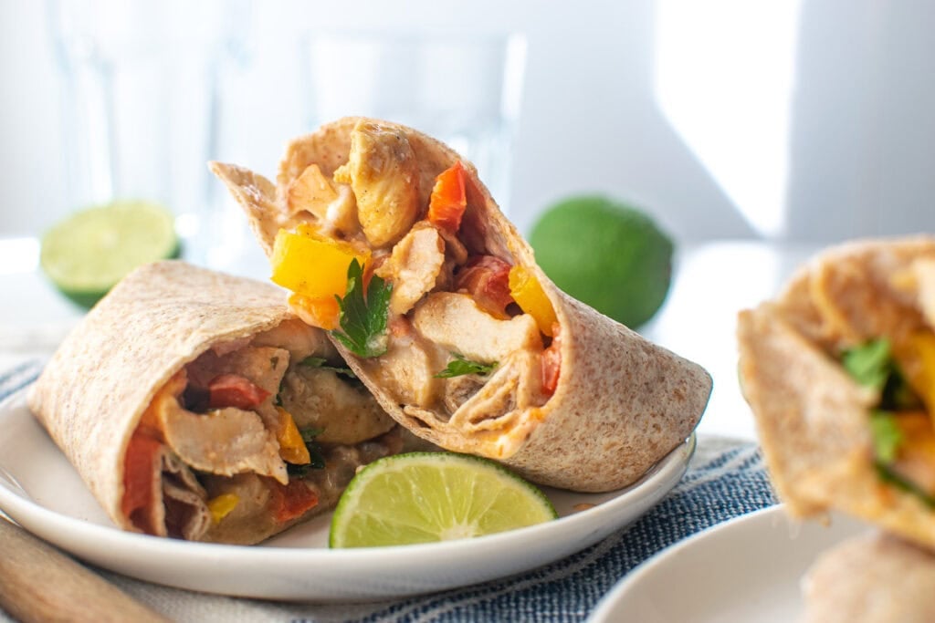 A chicken fajita in a tortilla wrap cut in half to show the filling on a white plate with a lime wedge.