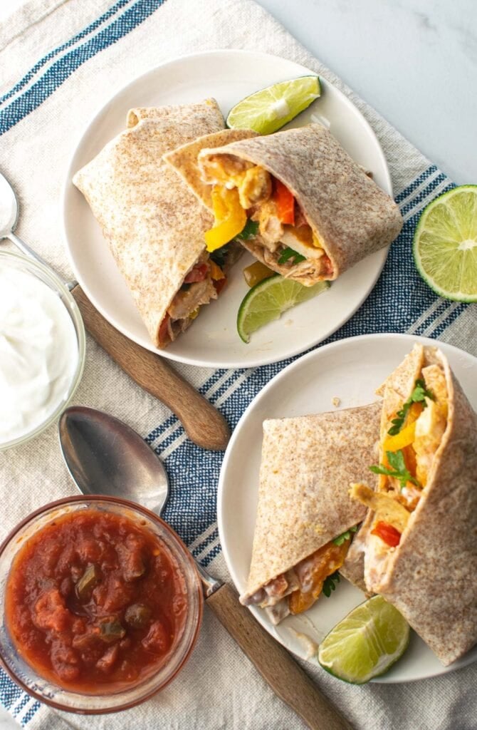 Two chicken fajitas on white plates sitting on top of a napkin with a blue stripe and bowls of salsa and sour cream to the side.