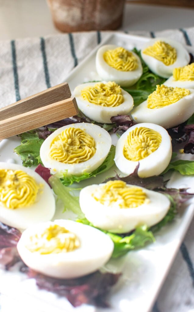 A platter of dill deviled eggs on a bed of lettuce with wooden tongs beside them on the platter.