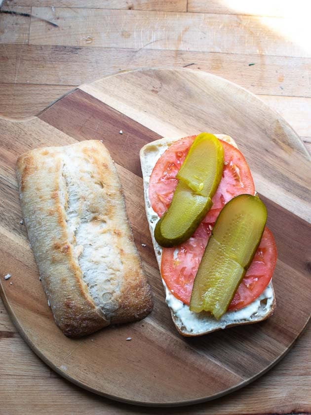 Dill pickle slices layered on top of sliced tomato and tarter sauce on the bottom half of a split ciabatta bun.