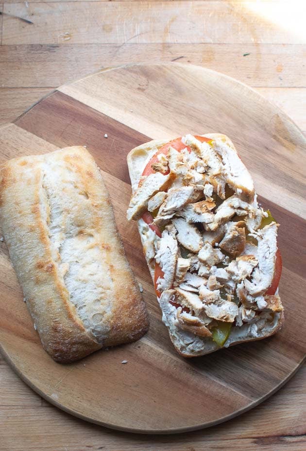 Sliced cooked chicken breast arranged over dill pickle slices, tomato slices and tartar sauce on the bottom of a split ciabatta bun.