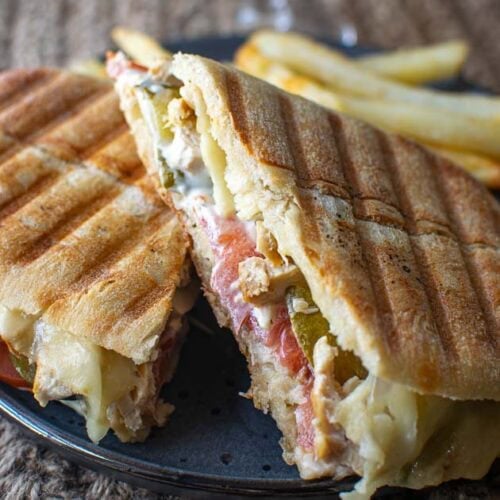 A chicken panini sandwich cut into two halves showing the filling of melted cheese over top of sliced chicken breast pieces, sliced tomatoes, dill pickles and tarter sauce.
