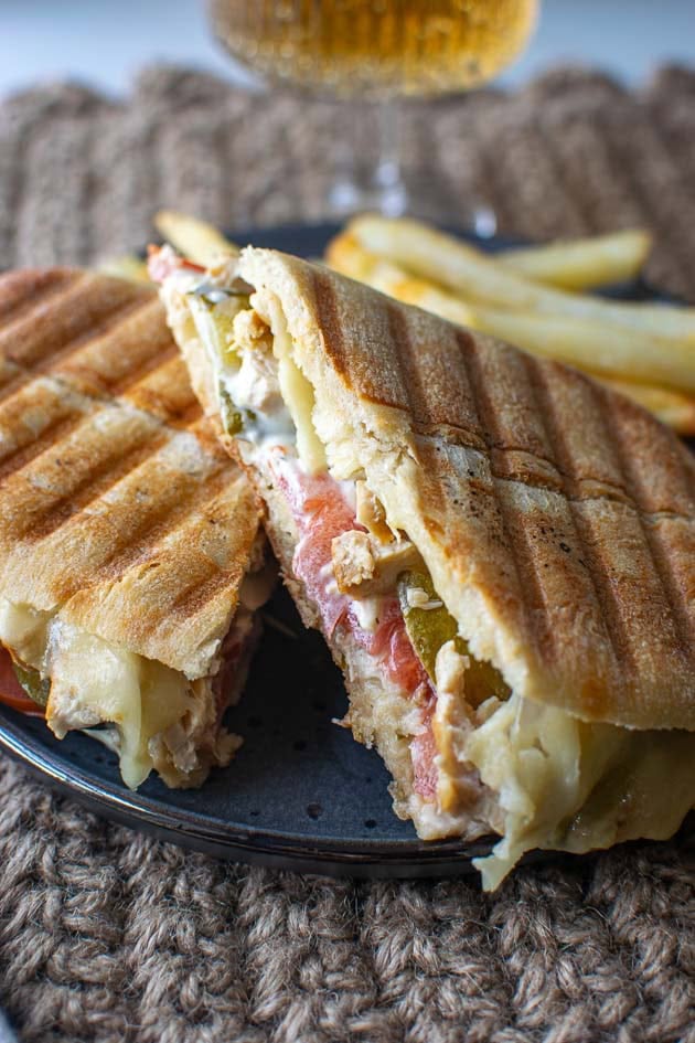 A chicken panini sandwich cut into two halves showing the filling of melted cheese over top of sliced chicken breast pieces, sliced tomatoes, dill pickles and tarter sauce.