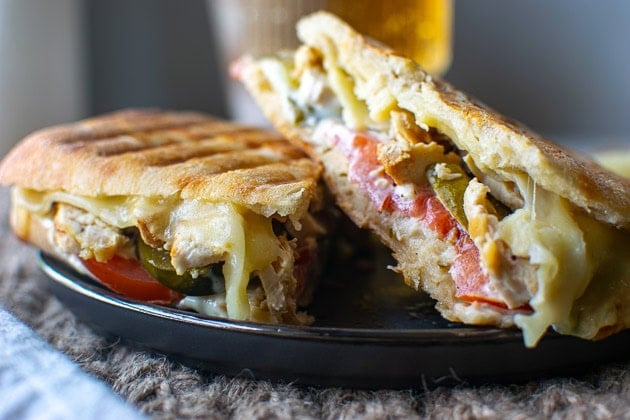 Chicken panini sandwich filled with tomatoes, pickles, cheese and tarter sauce on a black plate.