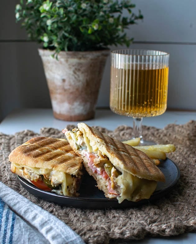 A chicken panini sandwich cut in half on a black plate with a side of French fries and a stemmed glass of ginger ale in the background.
