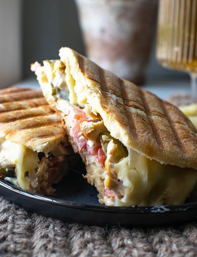 Chicken Panini Sandwich filled with chicken, melted cheese, tomatoes and dill pickles, served on a black plate.