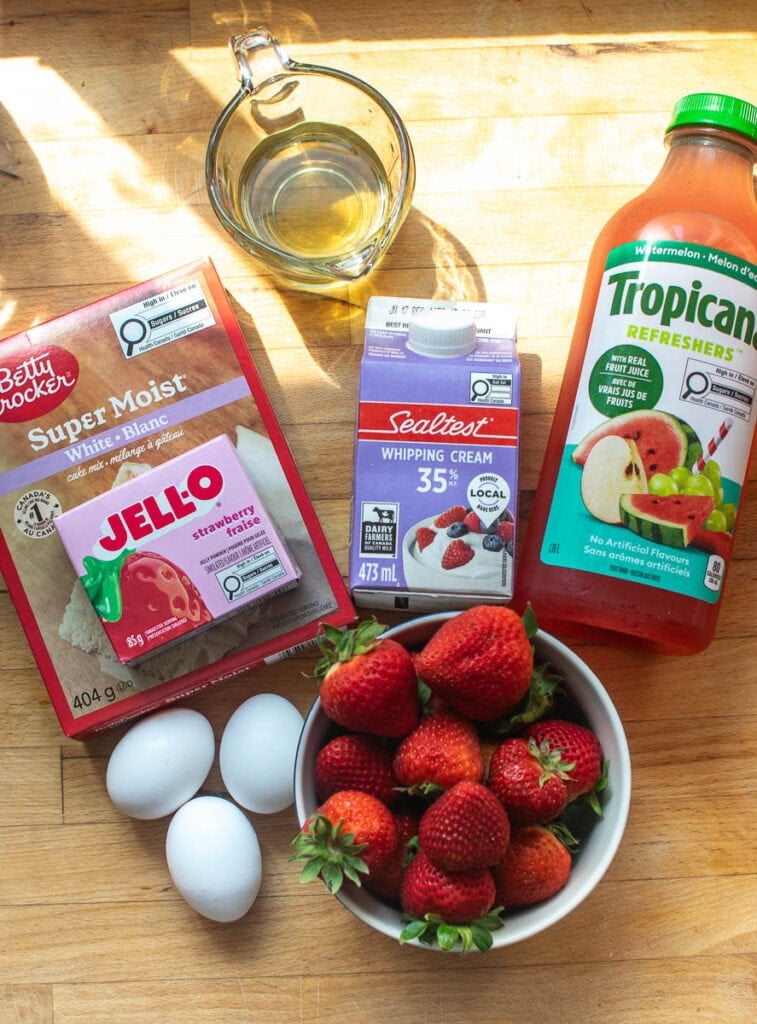 Ingredients to make watermelon layer cake with strawberry whipped cream frosting including Tropicana Watermelon juice, strawberry jello powder, whipping cream, eggs, oil, a cake mix, and fresh strawberries.
