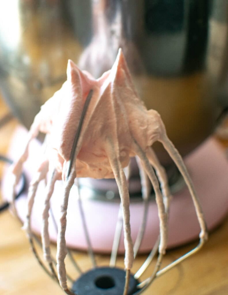 A whisk attachment upside down showing strawberry whipped cream whipped to stiff peaks.