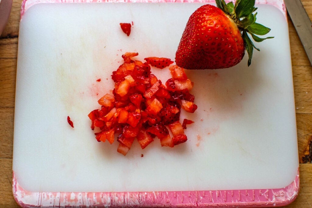 A full strawberry and some finely chopped strawberry pieces on a white cutting board.