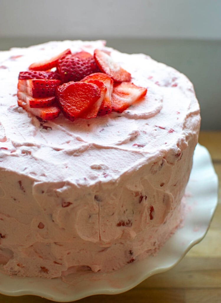 A watermelon flavoured layer cake frosted with strawberry whipped cream and garnished with sliced strawberries sitting on a ruffled cake plate.
