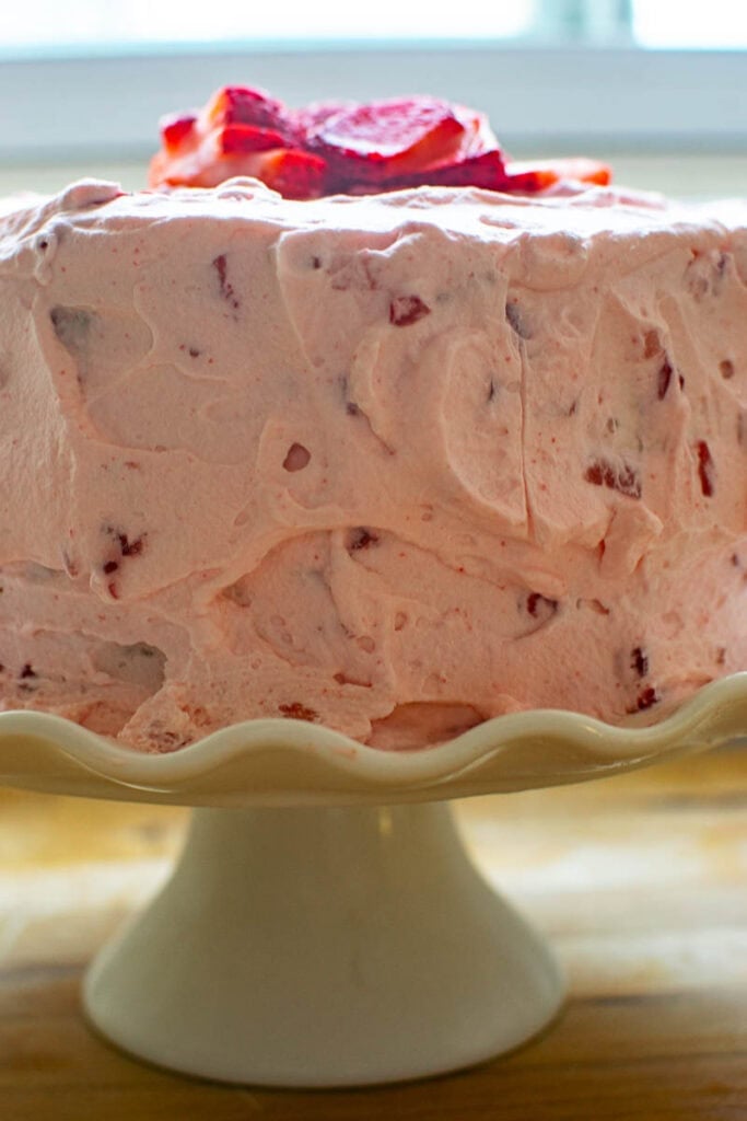 A watermelon layer cake frosted with fresh whipped cream with strawberries and garnished with slicked strawberries, on top of a white ruffled cake plate.