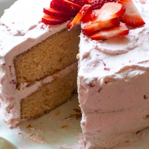 A watermelon layer cake with strawberry whipped cream frosting with a slice out on a white cake plate with ruffled edges.