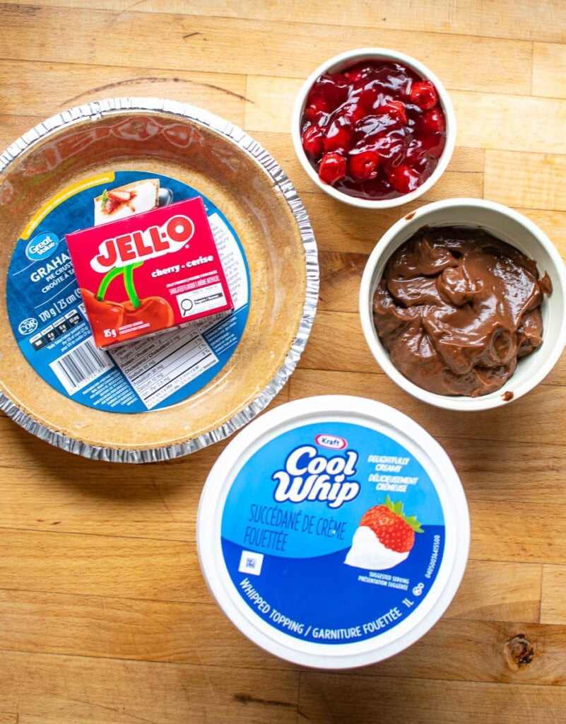 Ingredients to make a no-bake cherry chocolate pie including graham cracker crust, Cool Whip, Jell-O powder and cherry pie filling.
