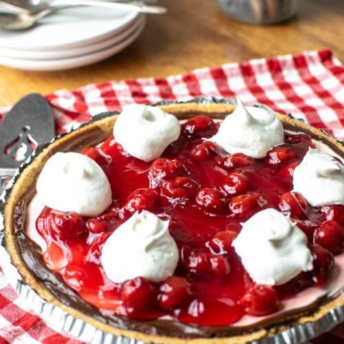 A no-bake chocolate cherry pie garnished with whipped cream sits on top of a red gingham napkins with a pie server and plates at the ready.