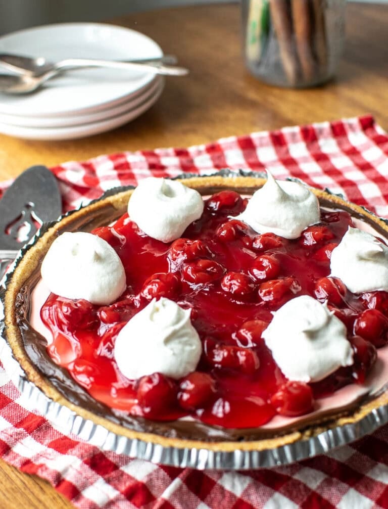 A no-bake chocolate cherry pie garnished with whipped cream sits on top of a red gingham napkins with a pie server and plates at the ready.