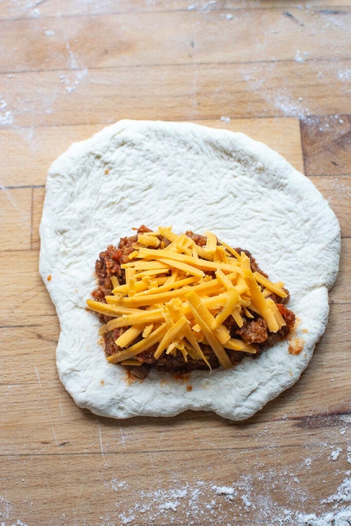 A ball of pizza dough rolled out into a rough circle with ground beef filling and shredded cheddar cheese on top.