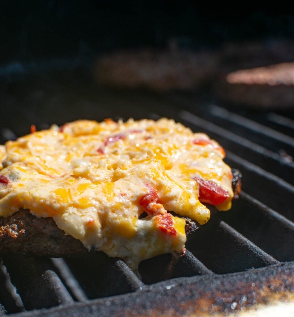 Pimento cheese spread spread on a grilled hamburger patty heating on the grill until melted.