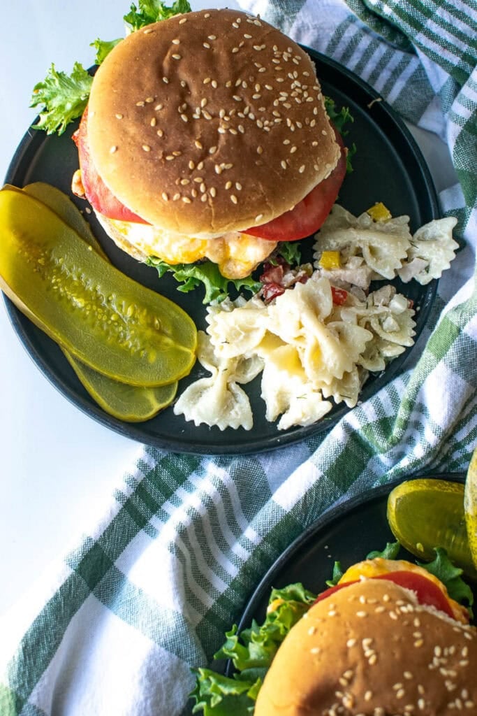 A pimento cheese burger on a black plate with a side of pickles and bowtie pasta salad.