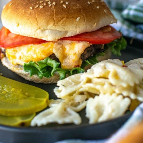 A grilled pimento cheese burger topped with lettuce and tomato on a plate with bowtie pasta salad.