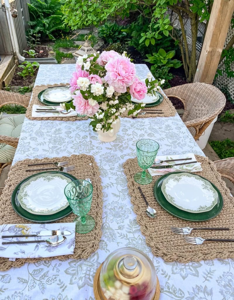 A table in the garden set for a summer dinner with a green floral tablecloth and green dishwater.