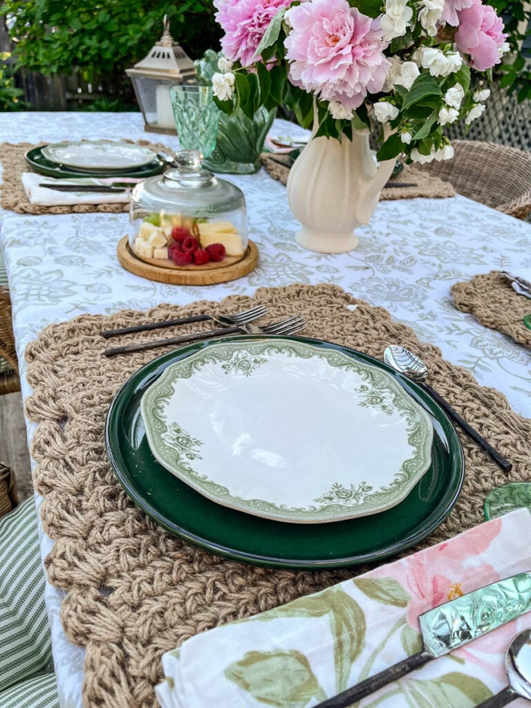 An outdoor table set for dinner with green dinner plates, rattan place mats, and a centrepiece of pink peonies and mock orange blossom branches.