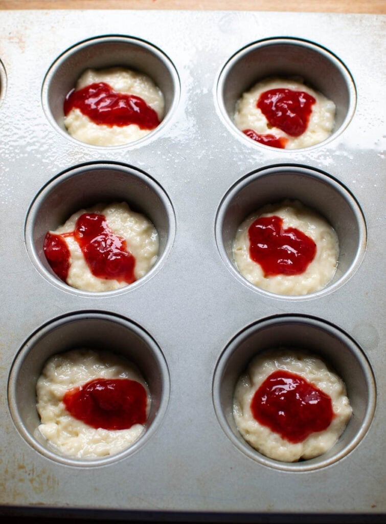 Muffin tins half filled with batter and topped with strawberry sauce.